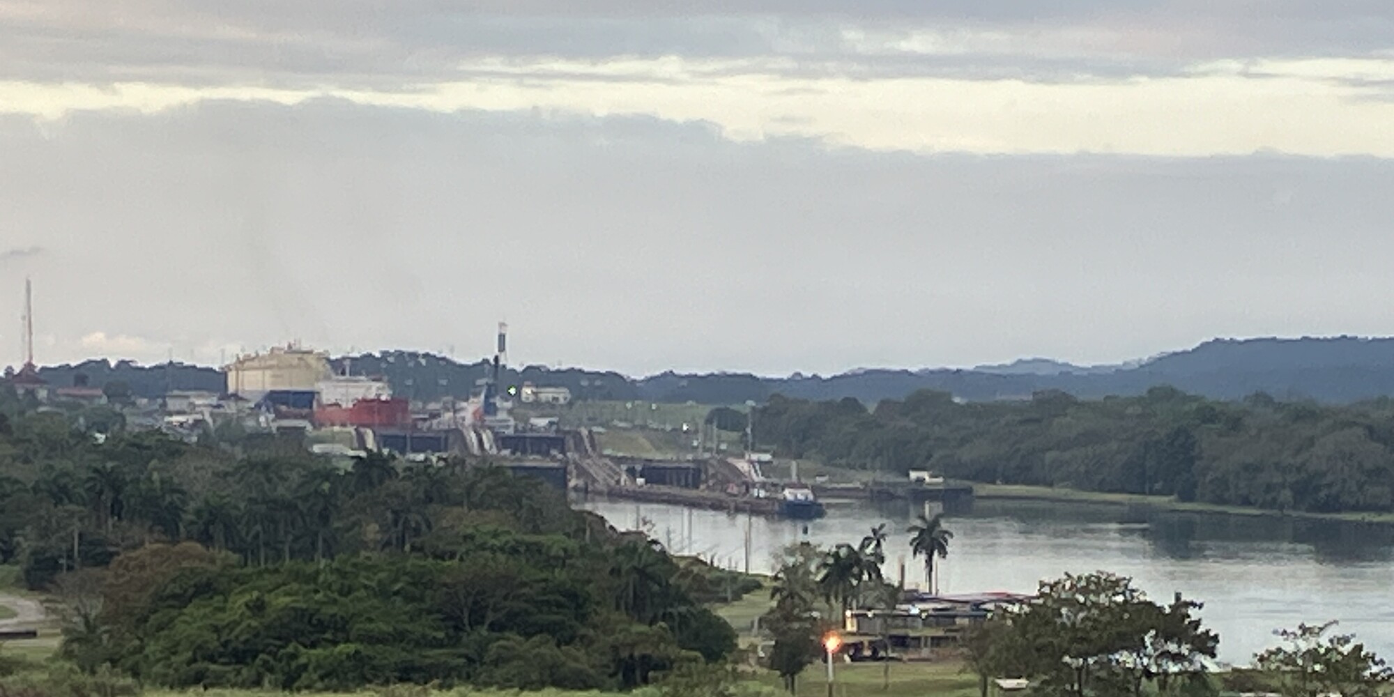 A broad lock chamber of the Panama Canal stretches through tropical hills under heavy clouds, with massive concrete walls and steel gates reflecting in still water